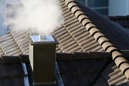 Close-up of a chimney with smoke rising above a roof with brown tiles.