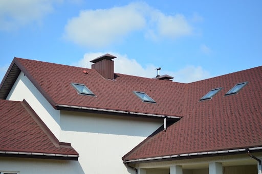 A close-up of a red-brown shingle roof with skylights and a chimney.